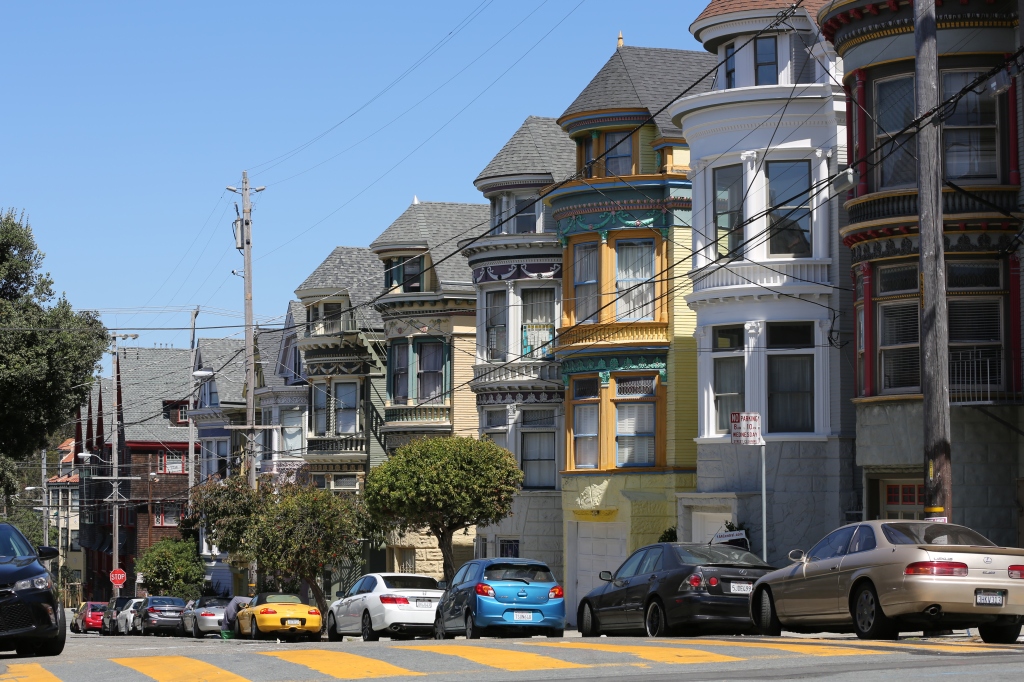 A row of Victorian homes in San Francisco with gasoline cars parked in front and power lines running down the street.